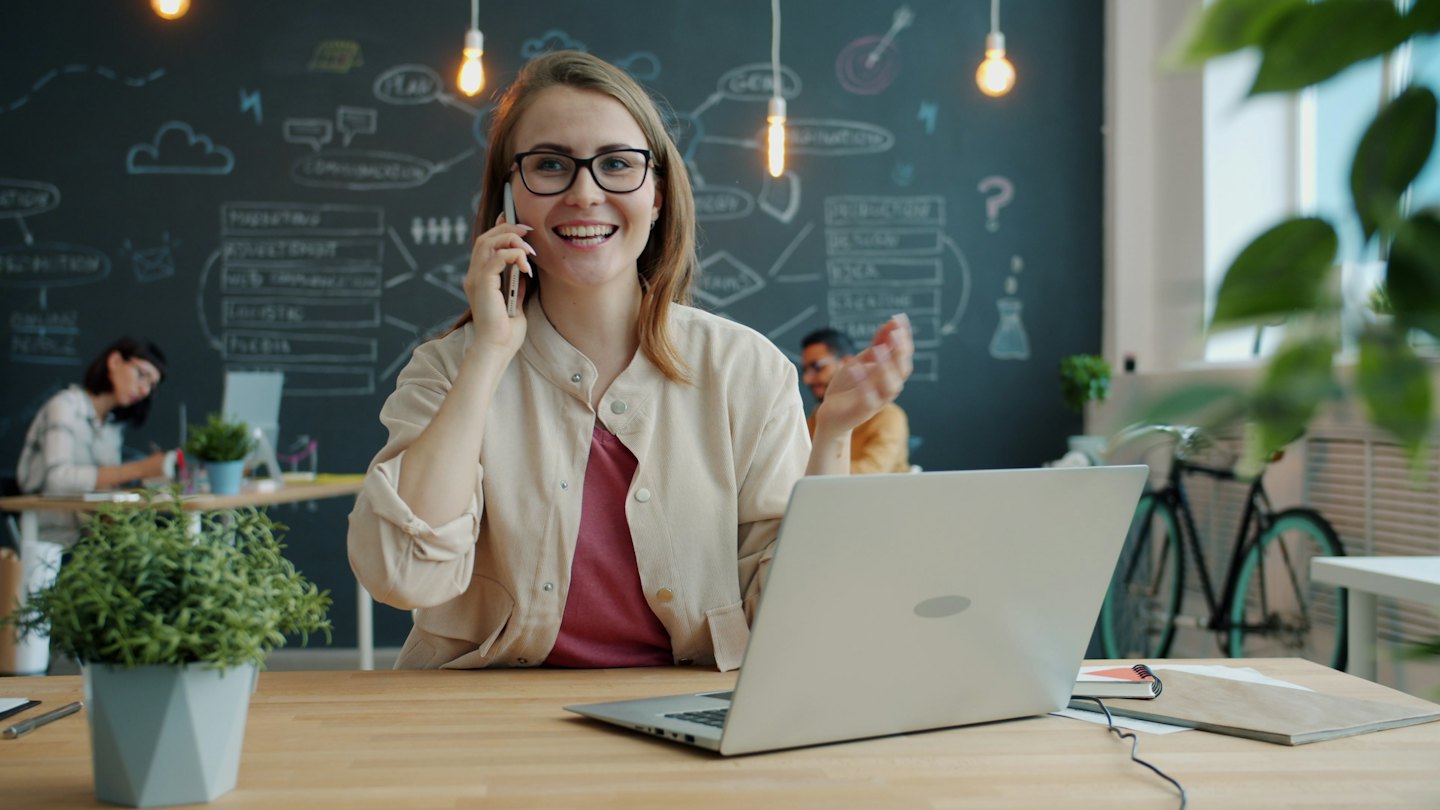 Mujer con gafas habla por teléfono en una oficina moderna.