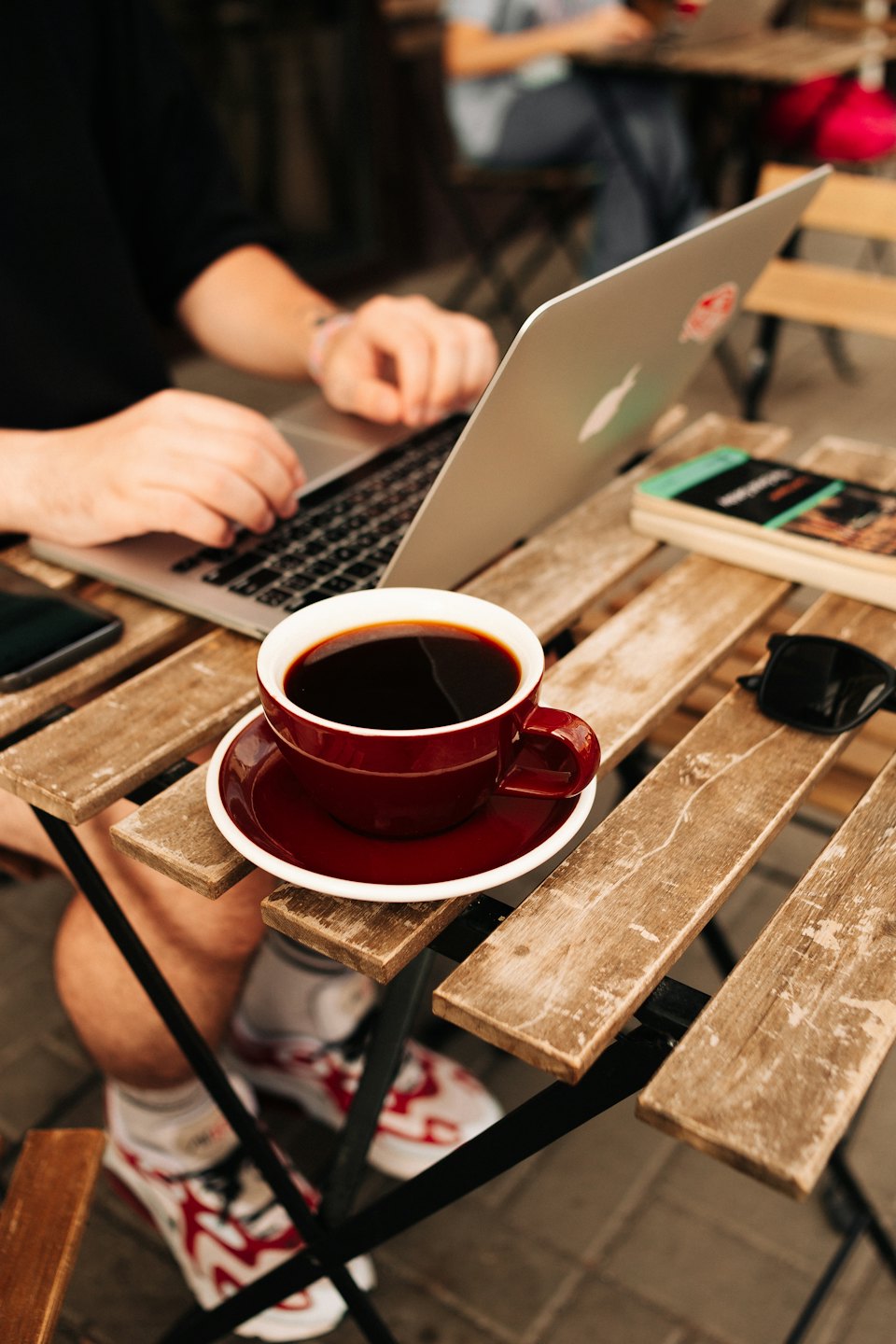 Manos escribiendo en un portátil plateado sobre la mesa de un café al aire libre.